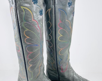 May include: A pair of gray leather cowboy boots with colorful embroidered floral and butterfly designs. The boots have a pointed toe, a mid-height heel, and are knee-high. The embroidery features rainbow colors.