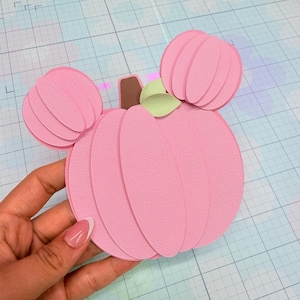 Puede incluir: Una manualidad de papel en capas con forma de calabaza con dos formas circulares más pequeñas unidas en la parte superior, que parecen orejas. La calabaza es de color rosa claro con un tallo marrón y una pequeña hoja verde claro texturizada.
