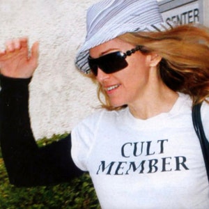 May include: A woman wearing a white hat with black stripes, sunglasses, and a white t-shirt with the text "CULT MEMBER" printed in black.