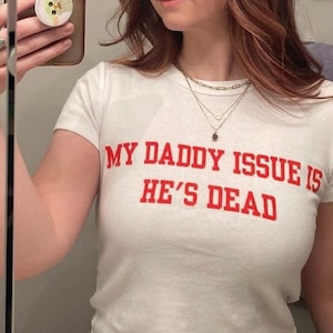 Puede incluir: Camiseta blanca con texto rojo que dice "MY DADDY ISSUE IS HE'S DEAD".