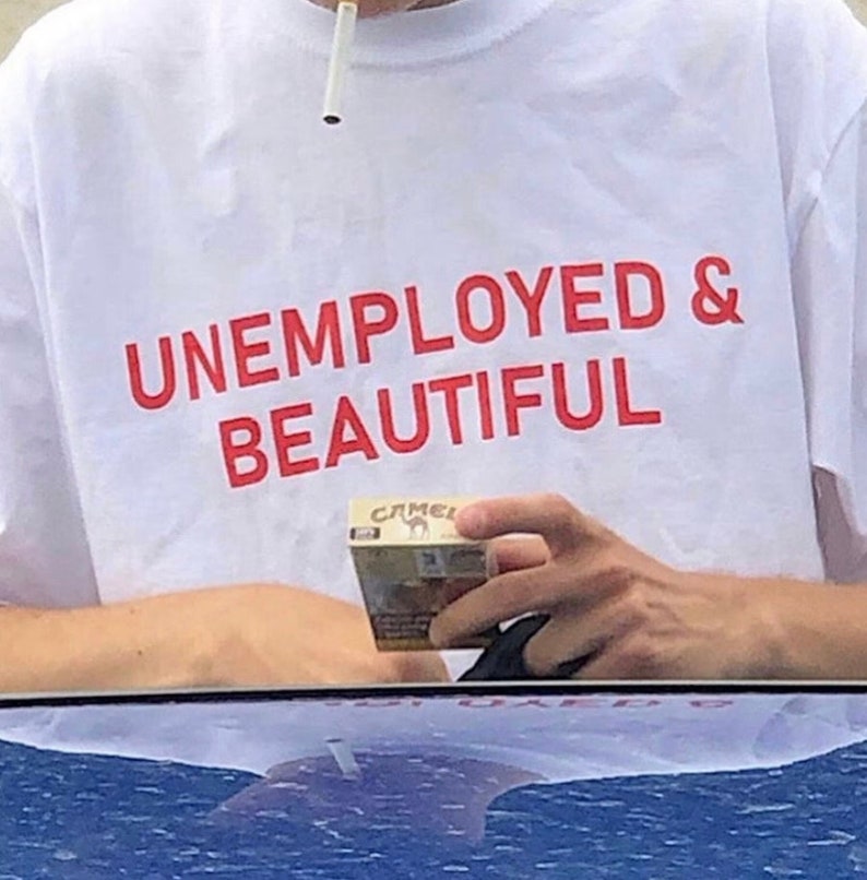 K&ouml;nnte beinhalten: Wei&szlig;es T-Shirt mit dem Text "UNEMPLOYED & BEAUTIFUL" in Rot.