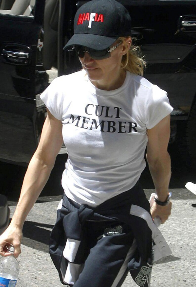 May include: A woman wearing a white t-shirt with the text "CULT MEMBER" printed on it. She is also wearing a black baseball cap with the word "MAFIA" printed in red letters.