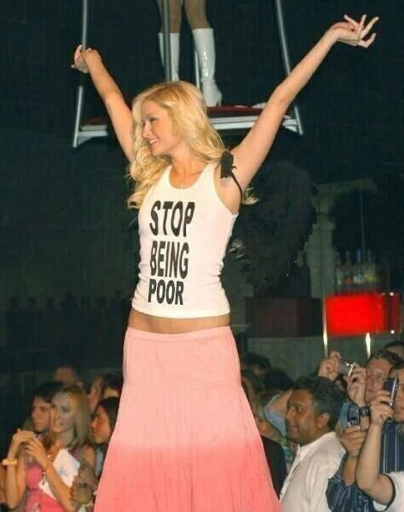 May include: A woman wearing a white tank top with the text "STOP BEING POOR" in black lettering. She is wearing a pink skirt and has her arms raised in the air.
