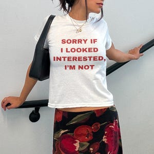 Pode incluir: Camiseta branca com o texto "SORRY IF I LOOKED INTERESTED, I'M NOT" impresso em vermelho.