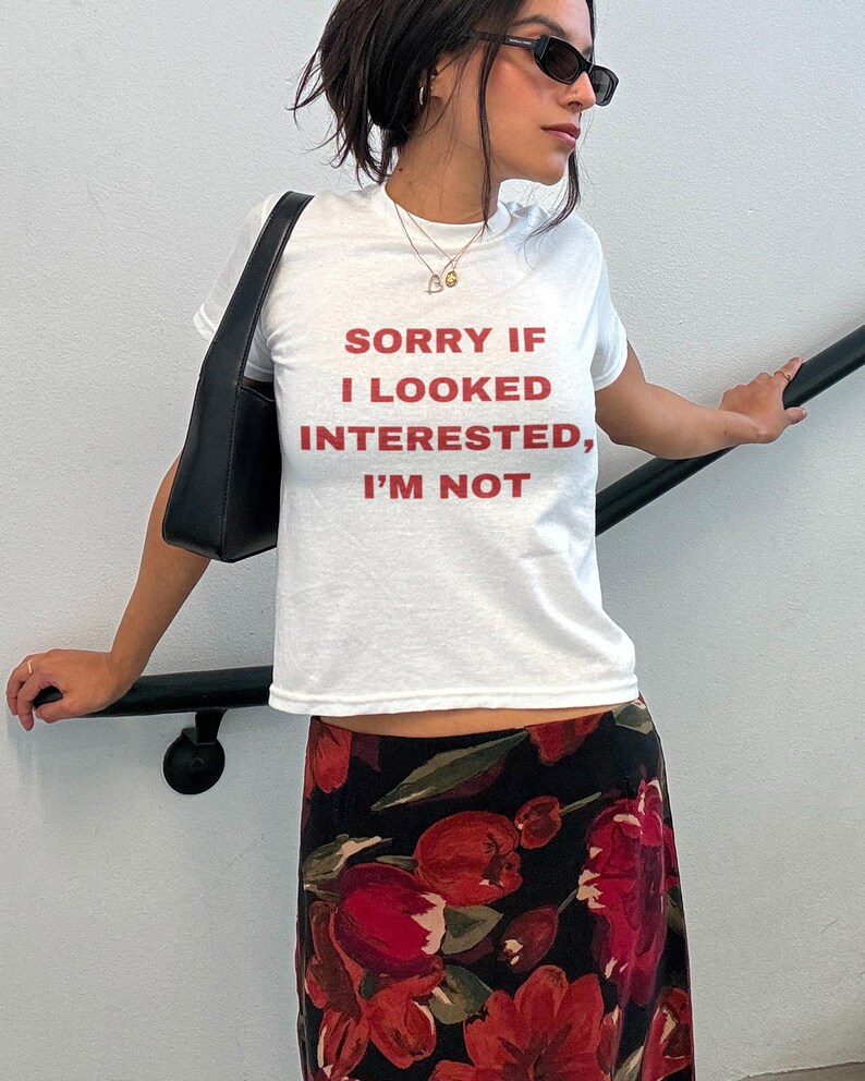 Pode incluir: Camiseta branca com o texto "SORRY IF I LOOKED INTERESTED, I'M NOT" impresso em vermelho.