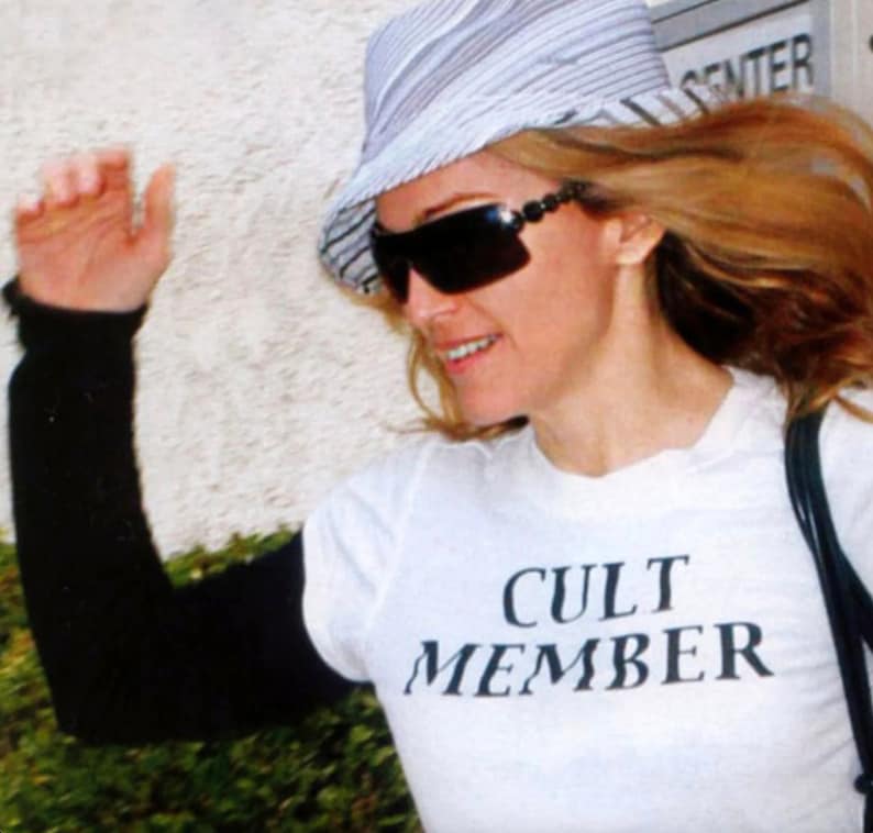May include: A woman wearing a white hat with black stripes, sunglasses, and a white t-shirt with the text "CULT MEMBER" printed in black.