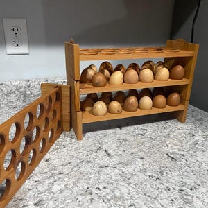 May include: Wooden egg rack with three shelves, holding several brown and beige eggs. A separate wooden egg holder with circular openings is on the left. The rack is made of light-colored wood and sits on a speckled countertop.