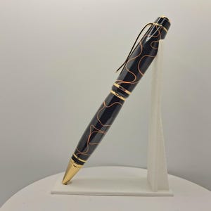 Op de afbeelding: Een zwarte balpen met een glanzende afwerking en dunne, golvende goudkleurige lijnen over de houder. De pen is voorzien van goudkleurige metalen accenten, waaronder een puntige tip, een centrale ring en een gebogen clip op de dop.