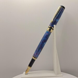 Op de afbeelding: Een vulpen met een blauw en paars gemarmerd ontwerp, gouden accenten en een gouden penpunt. De pen wordt weergegeven op een witte standaard, die het elegante ontwerp laat zien. De pen heeft een wervelend patroon.