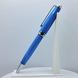 Op de afbeelding: Een blauwe en zilveren pen met een swirl-patroon op de cilinder. De pen staat op een witte standaard.