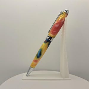 Op de afbeelding: Een balpen met een gemarmerde hars behuizing met wervelingen van geel, rood en blauw. De pen heeft een gepolijste chromen punt, een middenring en een zakclip. De pen staat schuin op een witte standaard om het kleurrijke patroon te tonen.