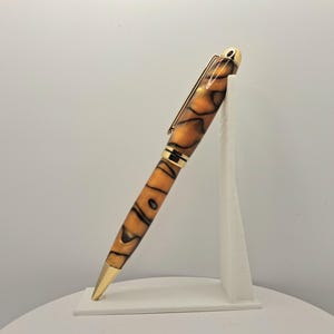 Op de afbeelding: Een balpen met een gemarmerde amberkleurige en zwarte hars behuizing en gepolijste goudkleurige metalen accenten, waaronder een clip, een middenring en een conische punt, schuin geplaatst op een witte standaard tegen een lichte achtergrond.