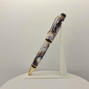 Op de afbeelding: Een pen van gemarmerde hars met goudkleurige accenten. De pen heeft een wervelend patroon van wit, zwart en bruin. De pen wordt gepresenteerd op een witte standaard tegen een witte achtergrond.