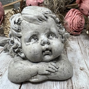 May include: A gray concrete cherub sculpture with wings, a cherubic face, and a sweet expression. The cherub is lying on its side with its arms crossed in front of its face.