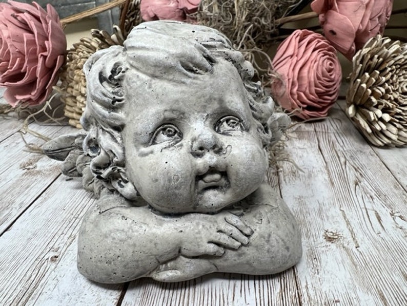 May include: A gray concrete cherub sculpture with wings, a cherubic face, and a sweet expression. The cherub is lying on its side with its arms crossed in front of its face.
