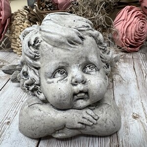 May include: A gray stone cherub sculpture with a sweet expression, resting its head on its hands. The cherub has curly hair and is looking to the side.