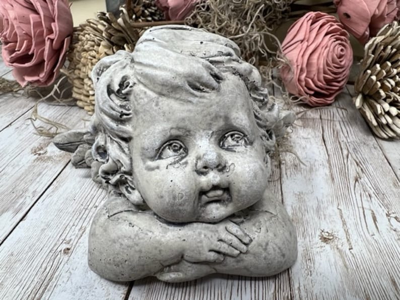 May include: A gray stone cherub sculpture with a sweet expression, resting its head on its hands. The cherub has curly hair and is looking to the side.