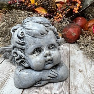 Puede incluir: Una estatua de querub&iacute;n gris con alas, descansando la cabeza en la mano. La estatua tiene una cara detallada con una expresi&oacute;n dulce y est&aacute; hecha de un material texturizado.