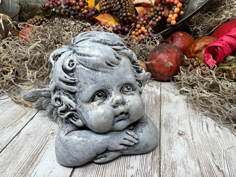 Puede incluir: Una estatua de querub&iacute;n gris con alas, descansando la cabeza en la mano. La estatua tiene una cara detallada con una expresi&oacute;n dulce y est&aacute; hecha de un material texturizado.