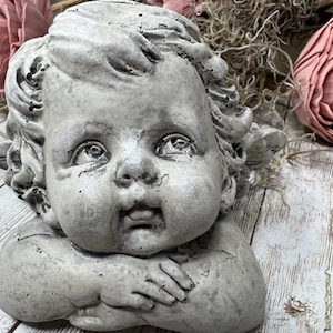 May include: A gray stone cherub statue with wings, the cherub is sitting with its arms crossed and has a sweet expression on its face.
