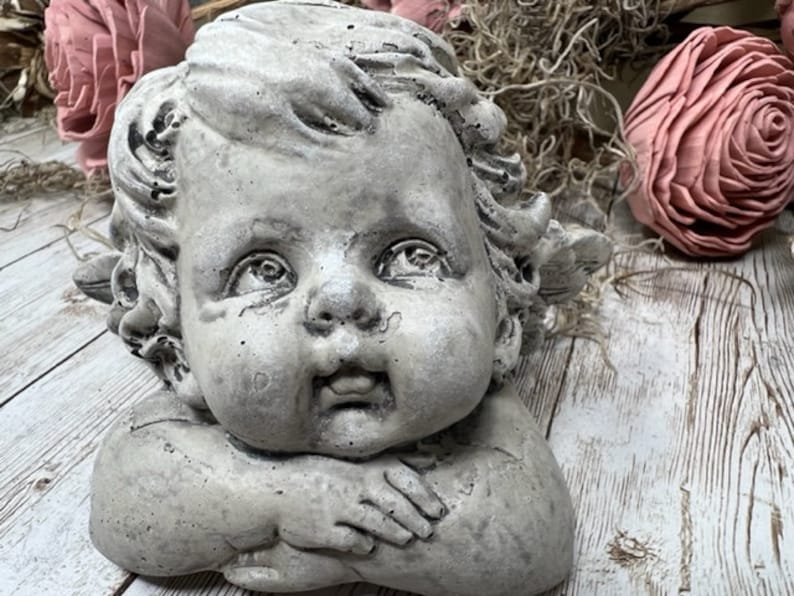May include: A gray stone cherub statue with wings, the cherub is sitting with its arms crossed and has a sweet expression on its face.