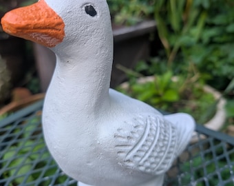 May include: A white goose figurine with an orange beak and black eye. The decorative goose has textured details on the wings and body. The goose is sitting on a green metal surface.