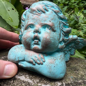 May include: A small, turquoise-colored cherub statue with wings. The cherub has a distressed, aged look with hints of copper. The cherub is holding its arms in front of its face.