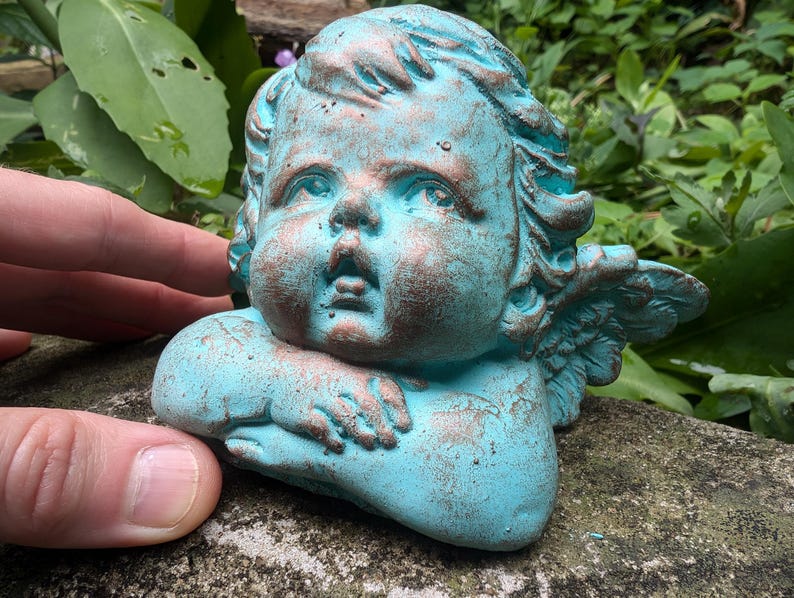 May include: A small, turquoise-colored cherub statue with wings. The cherub has a distressed, aged look with hints of copper. The cherub is holding its arms in front of its face.