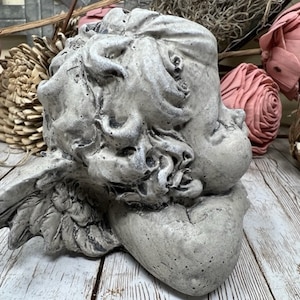 May include: A gray concrete cherub sculpture with wings, sleeping with its head resting on its arm. The sculpture is on a white wooden surface.