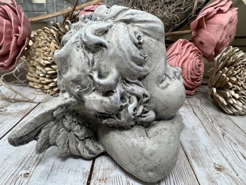 May include: A gray concrete cherub sculpture with wings, sleeping with its head resting on its arm. The sculpture is on a white wooden surface.