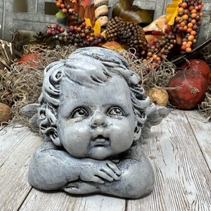Puede incluir: Una estatua de querub&iacute;n gris con alas, tumbada de lado con la cabeza apoyada en la mano. El querub&iacute;n tiene una expresi&oacute;n dulce en la cara y est&aacute; hecho de un material texturizado.
