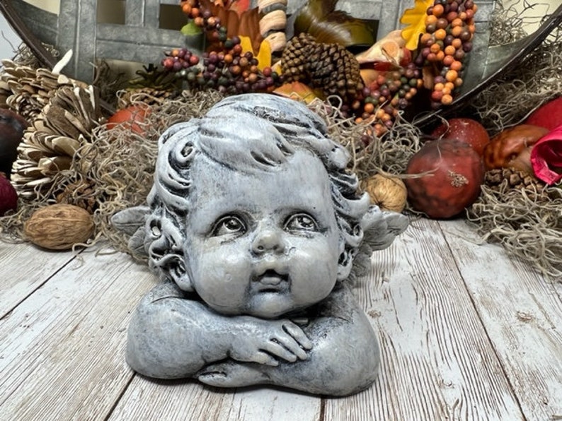 Puede incluir: Una estatua de querub&iacute;n gris con alas, tumbada de lado con la cabeza apoyada en la mano. El querub&iacute;n tiene una expresi&oacute;n dulce en la cara y est&aacute; hecho de un material texturizado.
