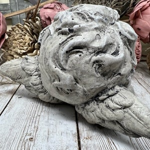 May include: A gray concrete sculpture of a cherub with wings, lying on its side. The sculpture has a weathered, distressed finish.