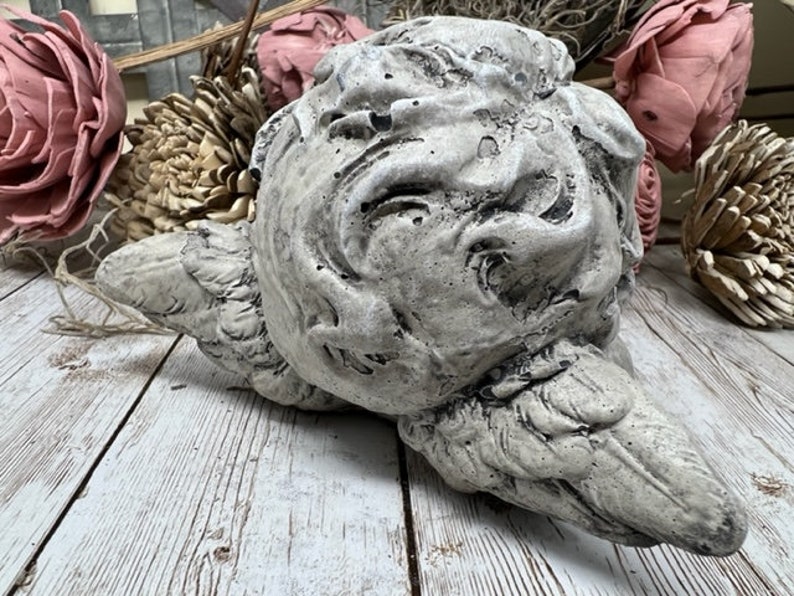 May include: A gray concrete sculpture of a cherub with wings, lying on its side. The sculpture has a weathered, distressed finish.