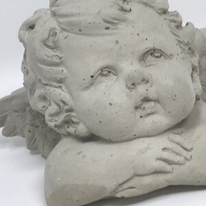 Puede incluir: Una escultura de querub&iacute;n de hormig&oacute;n gris con alas, descansando la cabeza sobre las manos. El querub&iacute;n tiene una dulce expresi&oacute;n en su rostro.