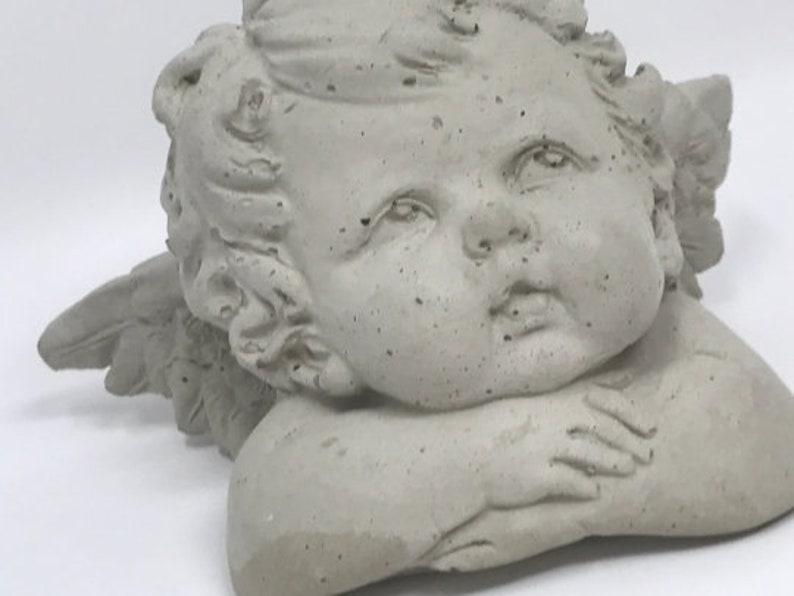 Puede incluir: Una escultura de querub&iacute;n de hormig&oacute;n gris con alas, descansando la cabeza sobre las manos. El querub&iacute;n tiene una dulce expresi&oacute;n en su rostro.