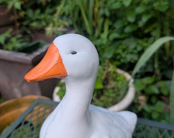 May include: A white ceramic goose figurine with an orange beak. The goose is perched on a green metal surface, with a blurred background of green foliage. The figurine has a smooth, matte finish.