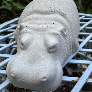 May include: A gray concrete hippopotamus sculpture. The sculpture is sitting on a blue metal table with a lattice design.