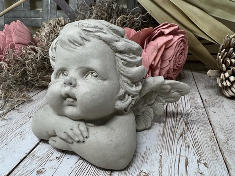 Puede incluir: Una estatua de querub&iacute;n de hormig&oacute;n gris con alas, mirando hacia arriba con una expresi&oacute;n dulce. El querub&iacute;n tiene una cara redonda con mejillas sonrosadas y tiene las manos juntas.