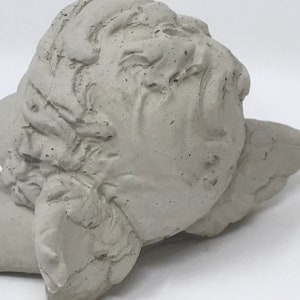 Puede incluir: Una escultura de hormig&oacute;n gris de la cabeza de un &aacute;ngel con alas. La cabeza del &aacute;ngel est&aacute; inclinada hacia un lado y las alas est&aacute;n extendidas detr&aacute;s de ella.