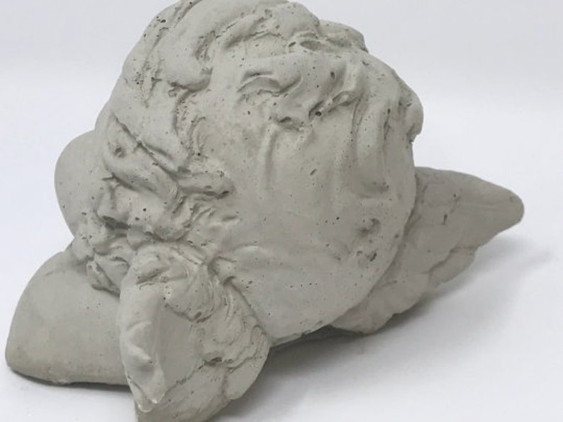 Puede incluir: Una escultura de hormig&oacute;n gris de la cabeza de un &aacute;ngel con alas. La cabeza del &aacute;ngel est&aacute; inclinada hacia un lado y las alas est&aacute;n extendidas detr&aacute;s de ella.