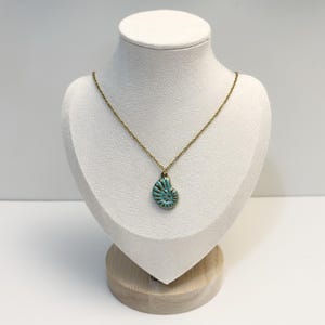 Könnte beinhalten: Eine Halskette mit einer goldfarbenen Kette und einem Anhänger in Form eines spiralförmigen Ammoniten-Fossils. Der Anhänger hat eine türkisfarbene Mitte mit goldfarbenen Rillen und Kanten und hängt an einer feinen Metallkette mit Gliedern.