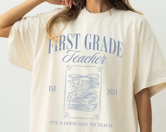 Può includere: Maglietta color avorio con una stampa grafica blu che dice "First Grade Teacher" con una pila di libri e il testo "EST. 2024" e "It's a good day to teach".