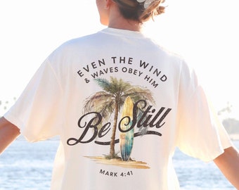 May include: White t-shirt with a palm tree and surfboard graphic. The text "Even the wind & waves obey him" is above the graphic and the text "Be Still" is below the graphic. The text "Mark 4:41" is at the bottom of the graphic.