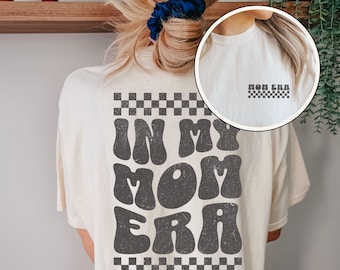 May include: A white t-shirt with a black checkered border and the text "In My Mom Era" in a retro, groovy font.