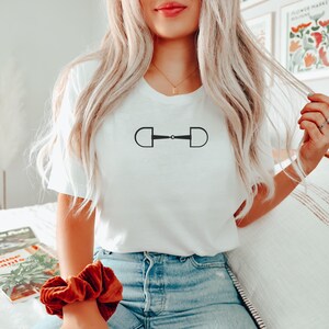 May include: A white t-shirt with a black equestrian bit design. The shirt is worn by a person with long blonde hair.