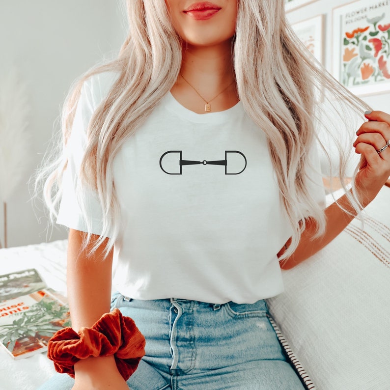 May include: A white t-shirt with a black equestrian bit design. The shirt is worn by a person with long blonde hair.