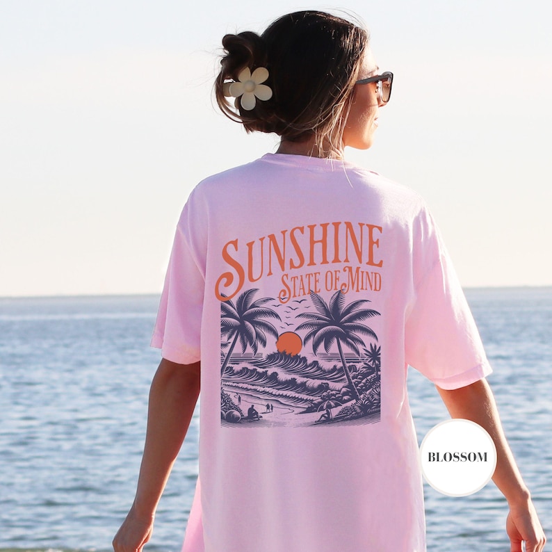 May include: A pink t-shirt with a graphic of a sunset over the ocean with palm trees and the text "Sunshine State of Mind".