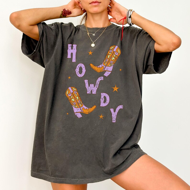 Puede incluir: Una camiseta gris oscuro de gran tama&ntilde;o con un estampado gr&aacute;fico de botas de vaquero moradas y la palabra "HOWDY" en letras moradas.
