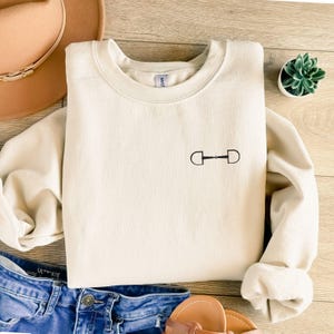 May include: A cream colored sweatshirt with a small black equestrian bit design on the front.